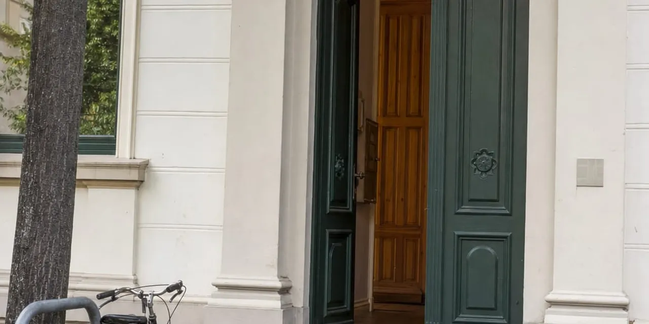 Geöffnete grüne Eingangstür eines hellen Altbaus mit Rundbogen, davor eine graue Rampe über die Stufen, links ein Baum und ein abgestelltes Fahrrad.